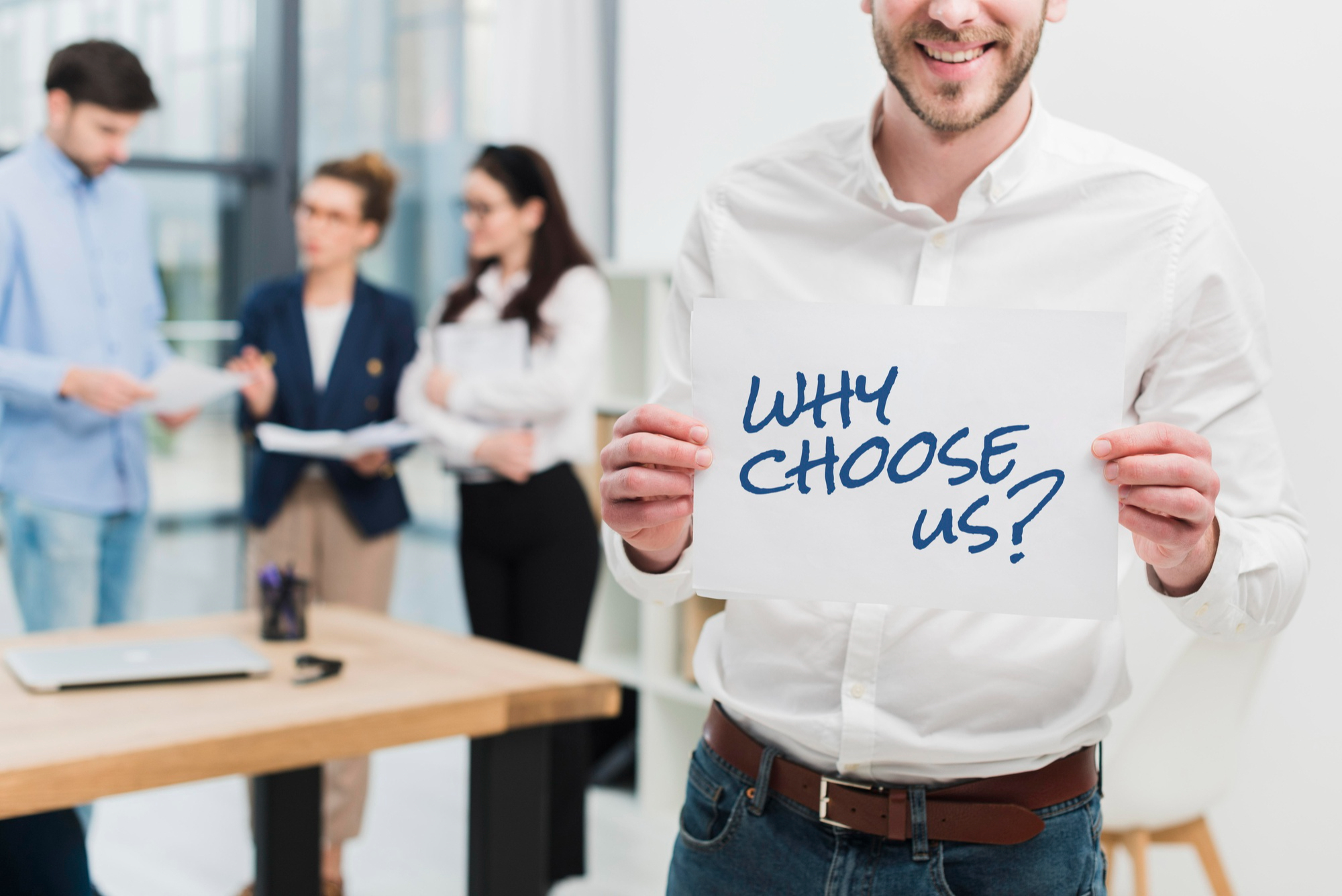 A smiling consultant holding a sign that reads ‘Why Choose Us?’ with a professional team discussing in the background, symbolizing expert guidance 
                        and trusted support for UK University Admission 2026 applicants.