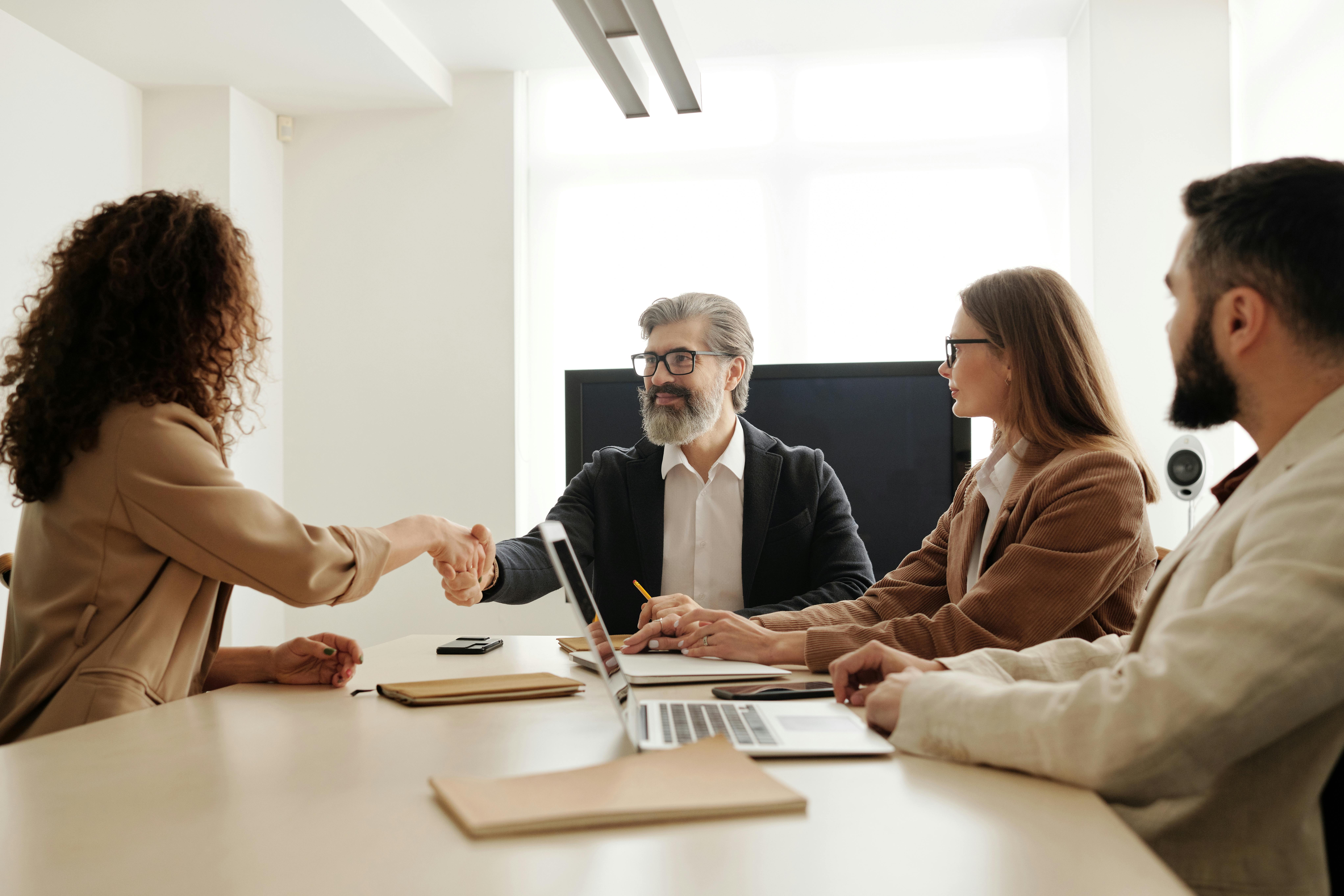 A professional consultation taking place in a bright office where a woman and a man shake hands across a desk, while two colleagues 
                        observe with laptops and notebooks open. The handshake symbolizes trust and partnership, reflecting the personalized guidance provided by UK 
                        University Admission Consultants to students seeking university placements in the UK.
