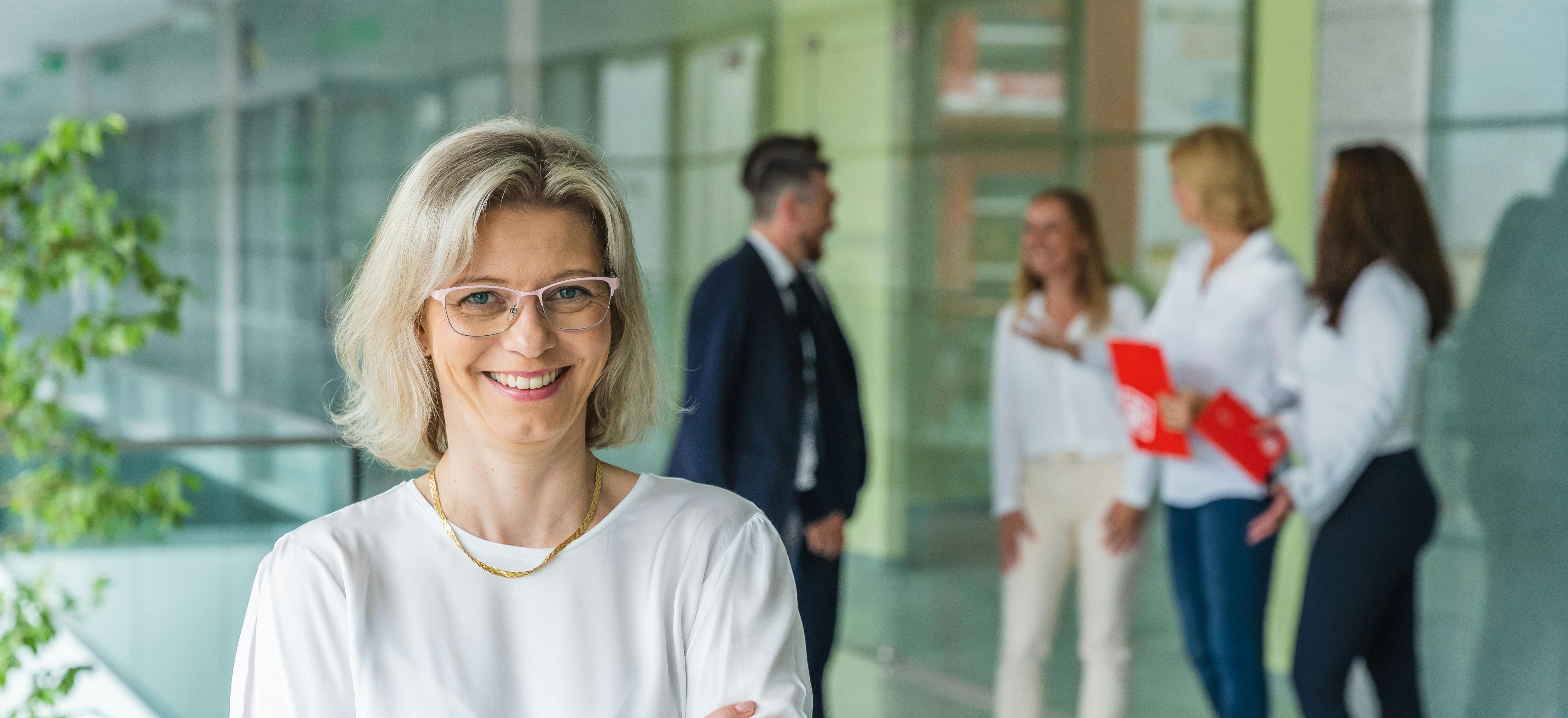 A confident professional woman with short blonde hair and glasses smiling with folded arms in a bright modern office, while a small team of colleagues 
                        converse in the background. The scene reflects the approachable and expert nature of UK University Admission Consultants offering comprehensive guidance to 
                        students pursuing higher education in the UK.